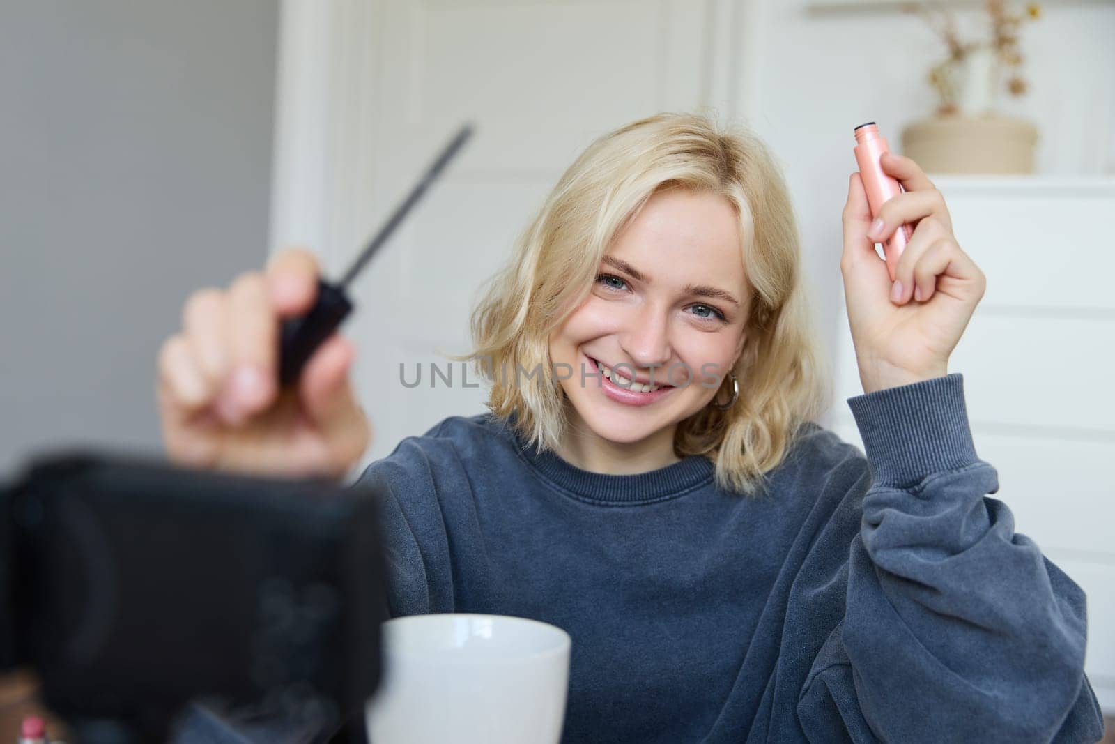 Portrait of young smiling woman in her room, recording video on camera, lifestyle vlog for social media, holding mascara, reviewing her makeup beauty products, showing how to use cosmetics.