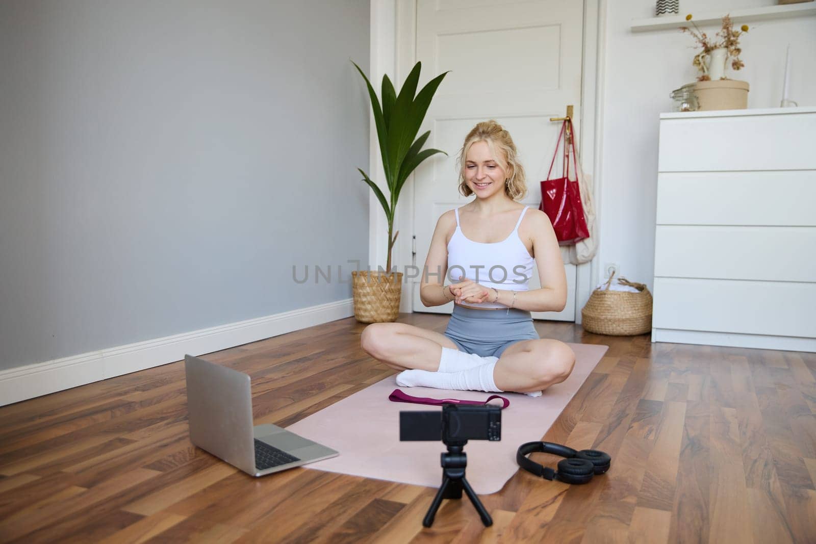 Portrait of young fitness blogger, woman showing exercises to her followers, recording video on digital camera, doing workout training session, home yoga, sitting in front of laptop on rubber mat.