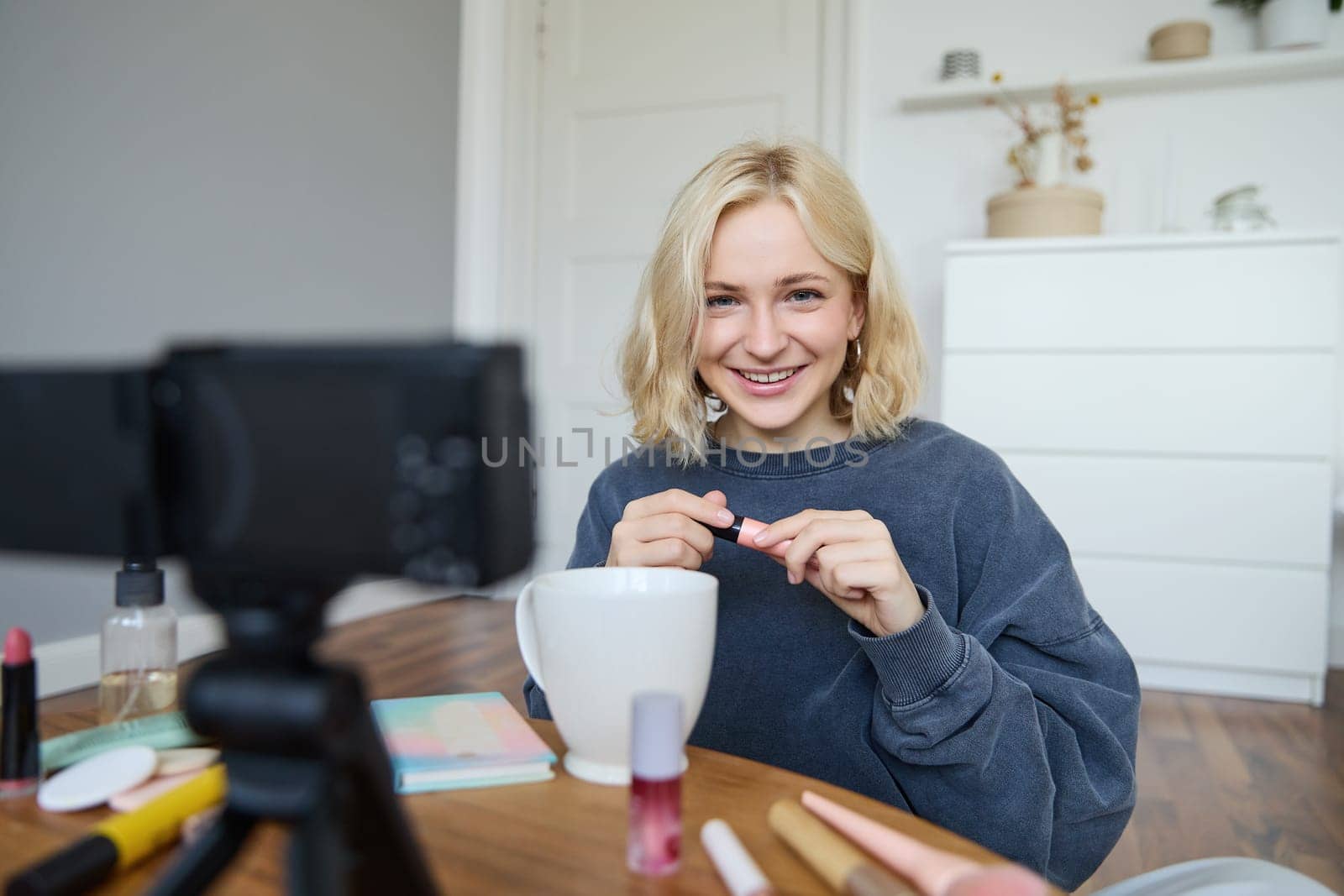 Portrait of blond smiling woman records a lifestyle blog, vlogger or makeup artist recording video for social media, holding mascara, reviewing beauty products for followers online.