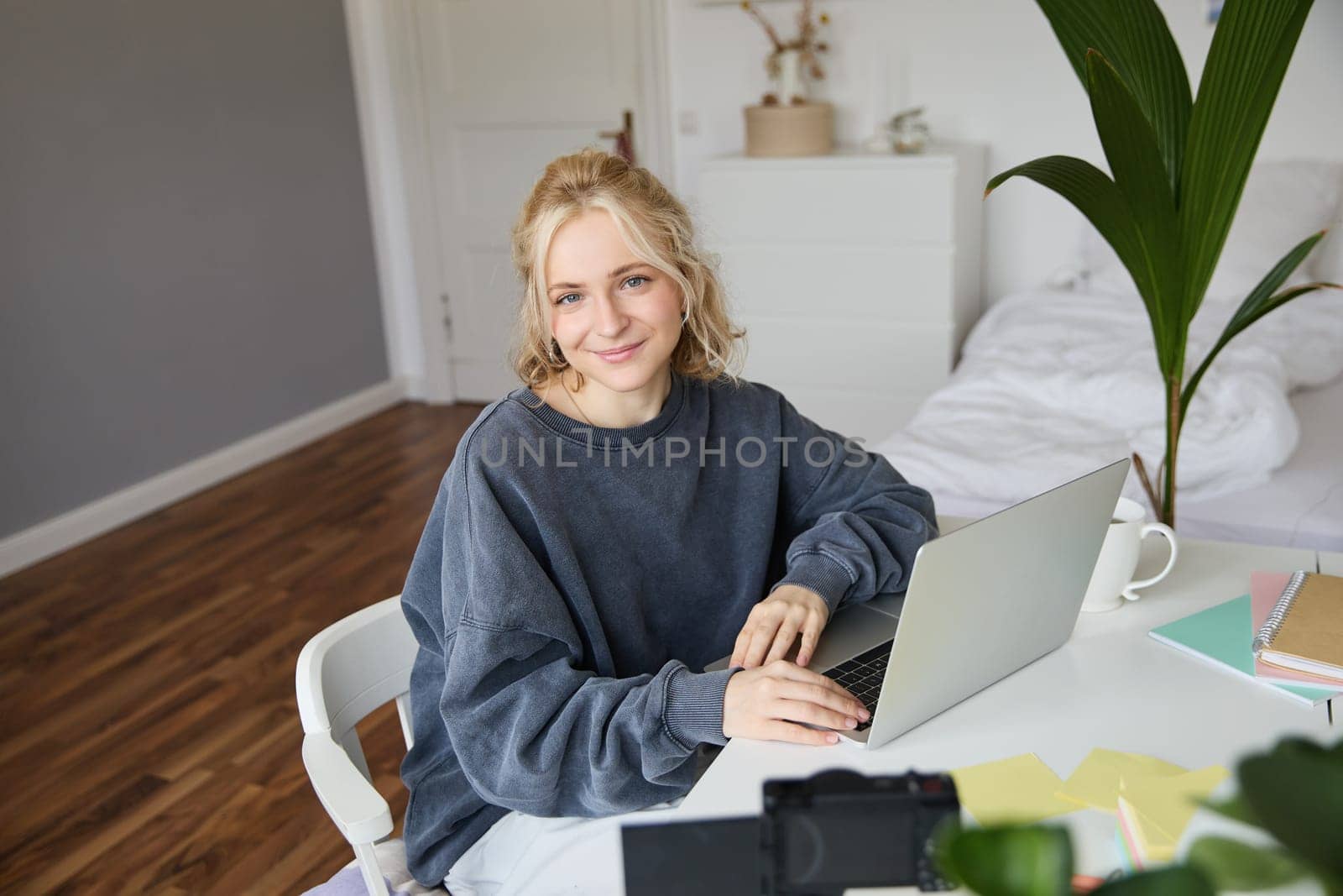 Portrait of young blond woman with laptop, recording video on digital camera, created video blog, vlogging in her room, editing on computer.