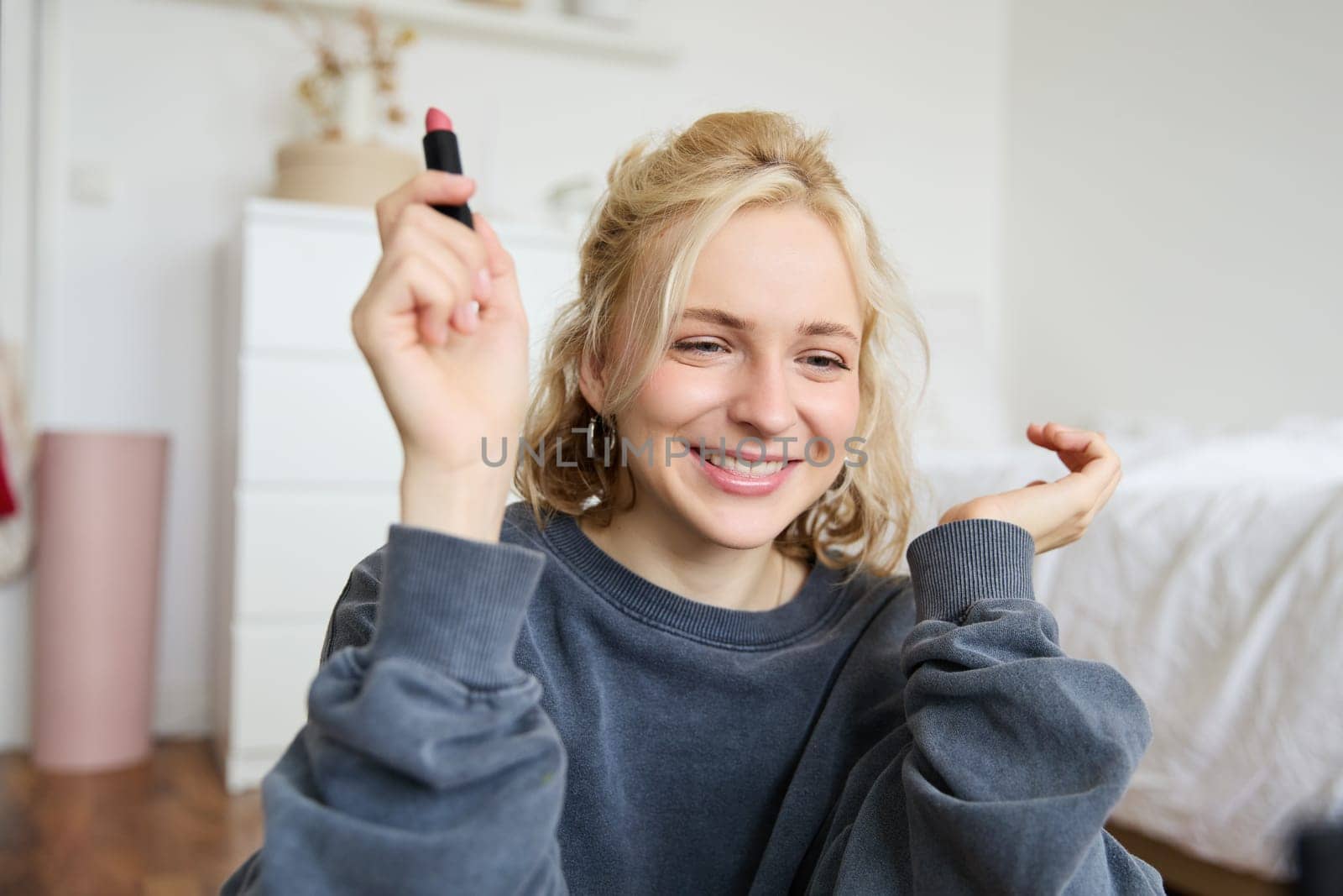 Portrait of young social media influencer, woman recording a video with beauty products, showing her makeup on camera, holding lipstick and smiling, sitting on floor.