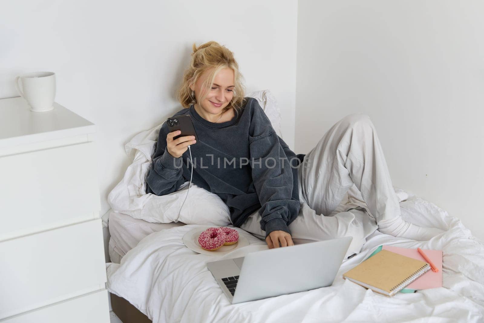 Lifestyle and weekend concept. Young smiling woman, relaxing at home, lying in bed with cup of tea and doughnut, using laptop and smartphone in bedroom.