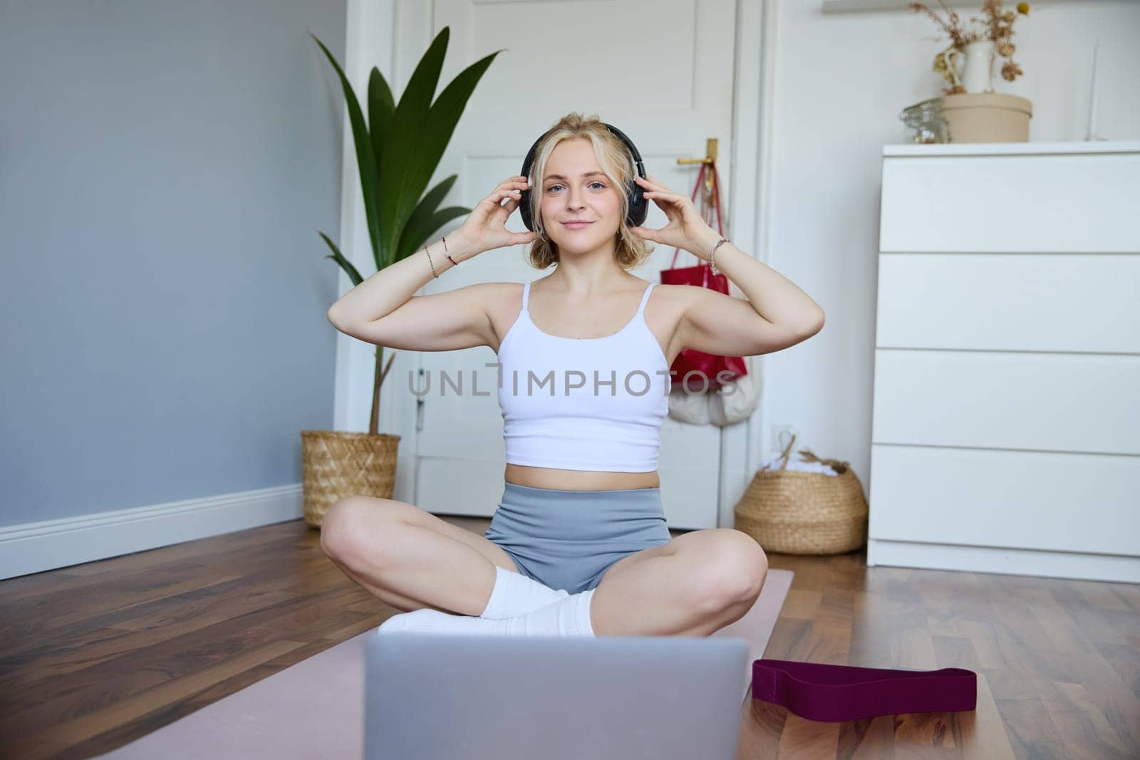 Portrait of young woman at home, connects to online gym training session, following fitness instructions on laptop, wearing wireless headphones during workout.