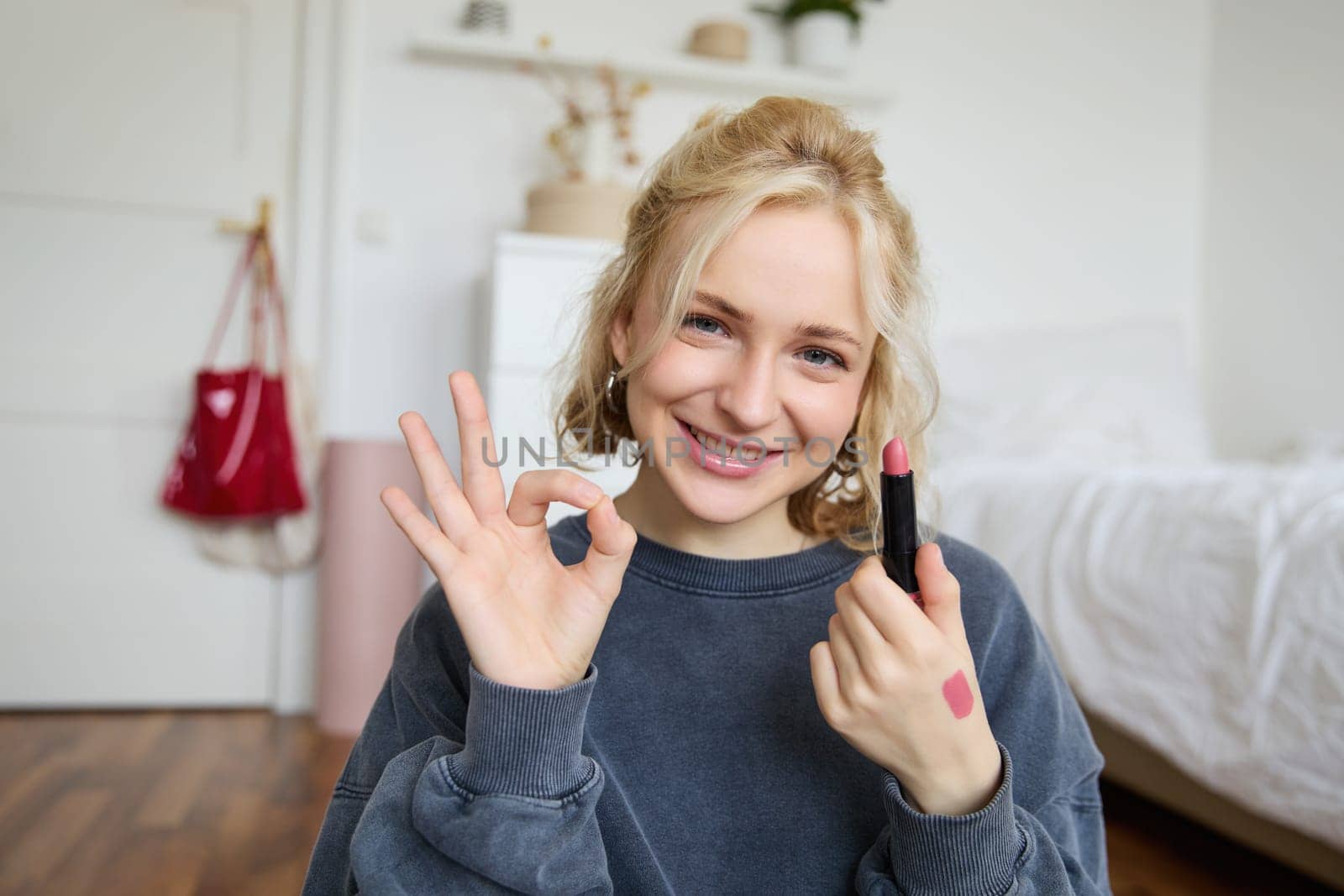 Portrait of young blond woman, content creator, recording video for social media about makeup, showing okay hand sign and lipstick, recommending good cosmetic product.