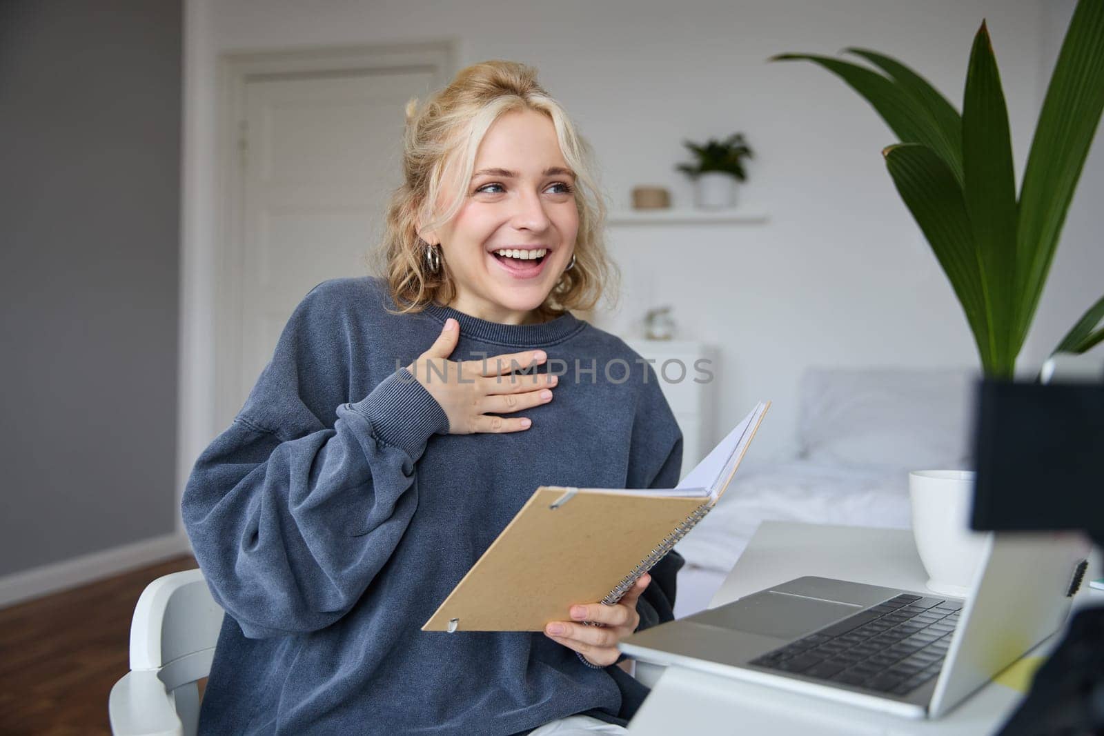 Portrait of smiling, beautiful young blond woman, student working on assignment from home, online learning in her bedroom, talking to video camera, chatting, holding notebook.