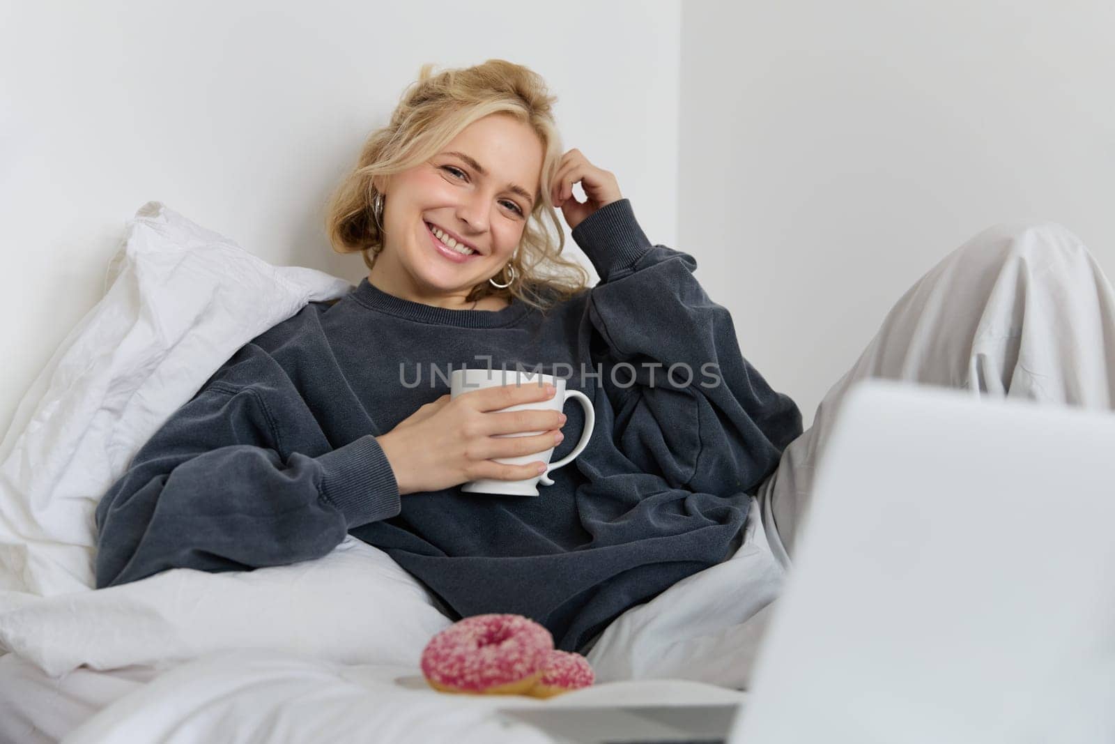 Portrait of happy blond girl, female model lying in bed in cosy clothes, eating and watching movie on laptop, smiling while looking at screen.