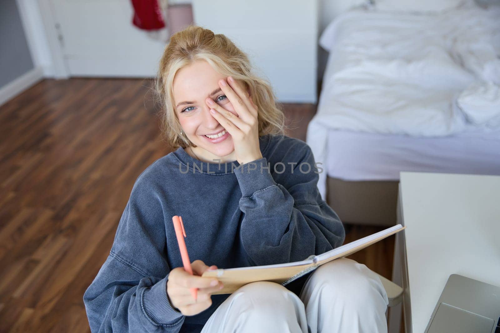 Portrait of smiling, charismatic young woman, writing down notes, making plans and putting it in planner, holding journal, sitting in bedroom and looking happy at camera.