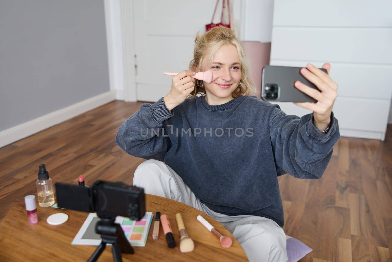 Portrait of beautiful blond girl doing makeup on camera, recording vlog using digital camera and live stream on mobile phone app, chatting with audience.