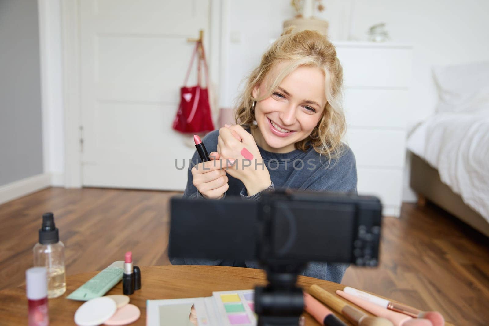 Image of young stylish woman, blogger recording a beauty lifestyle video of her picking best lipstick, showing lip balm swatches on her skin, sitting in front of digital camera in empty room.