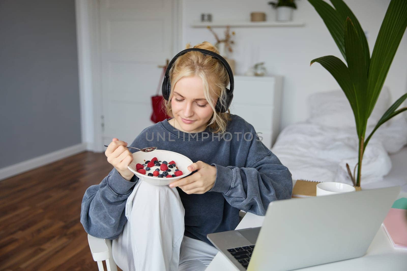 Portrait of smiling young blond woman in headphones, sitting in room, watching movie on laptop, eating breakfast and drinking tea, having lunch in front of computer screen.