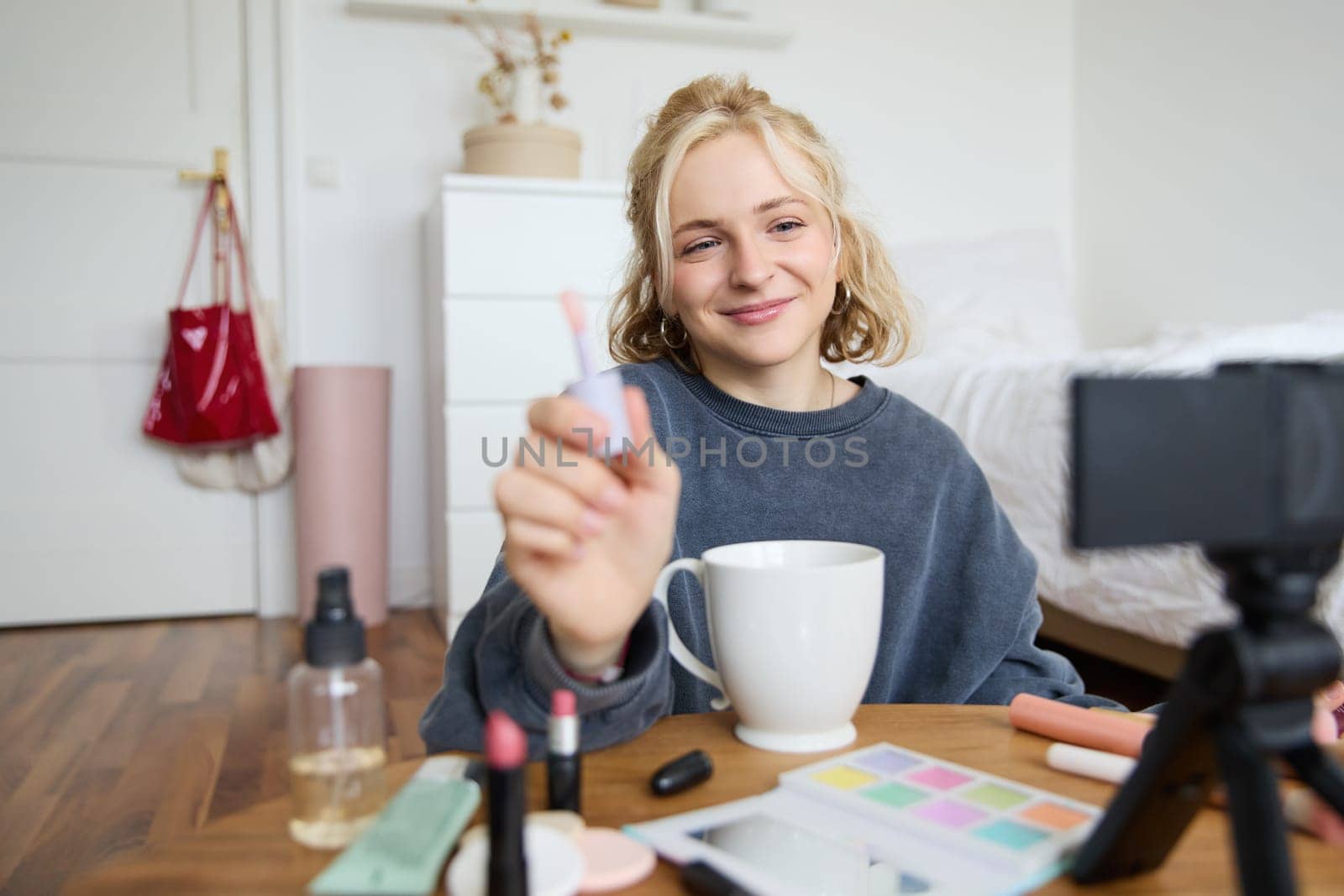 Lifestyle, beauty blogger, woman recording video of her putting on makeup, talking to camera, making online tutorial, showing her lip gloss or lipstick to followers, sitting on floor with cup of tea.