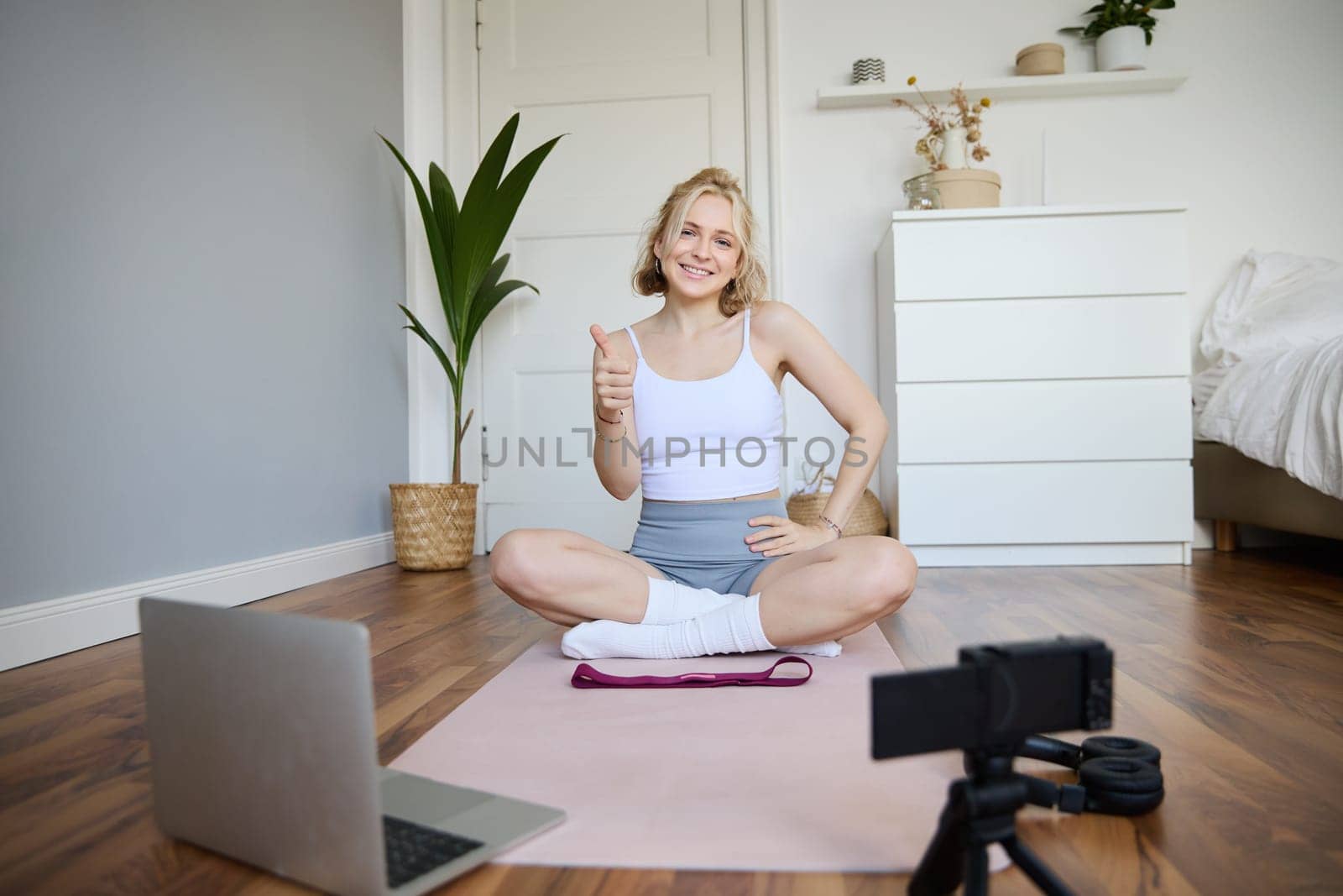 Young female athlete, fitness instructor woman sits on floor rubber mat, recording video on digital camera, showing how to workout, explaining exercises.