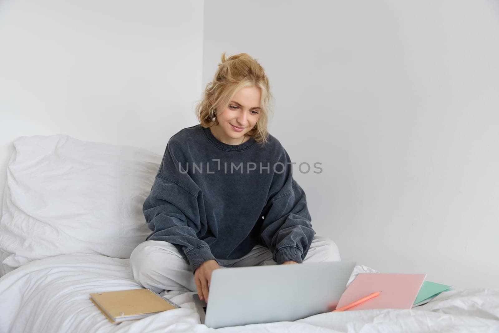 Image of happy young woman, student e-learning from home, connect to online course on her laptop, sits on bed with notebooks, smiling and looking happy.