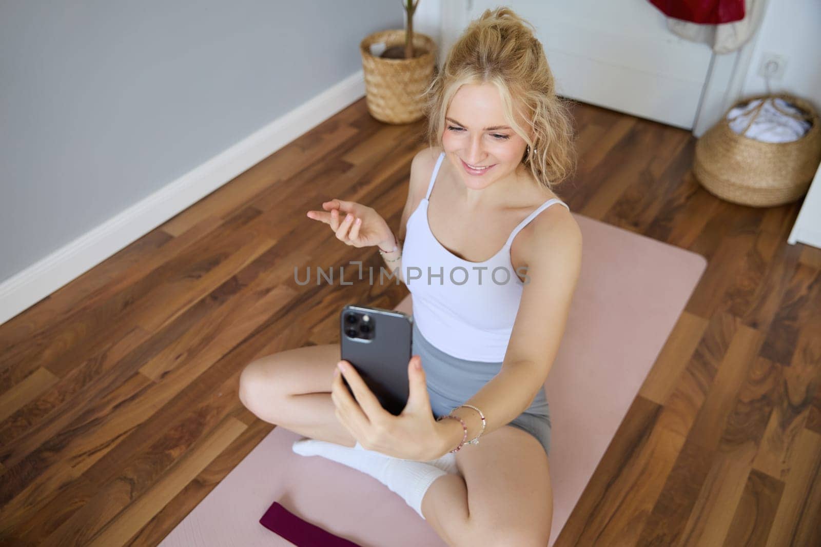 Portrait of young blogger, fitness instructor showing how to do exercises, sitting on yoga mat with smartphone, talking to followers on live stream.