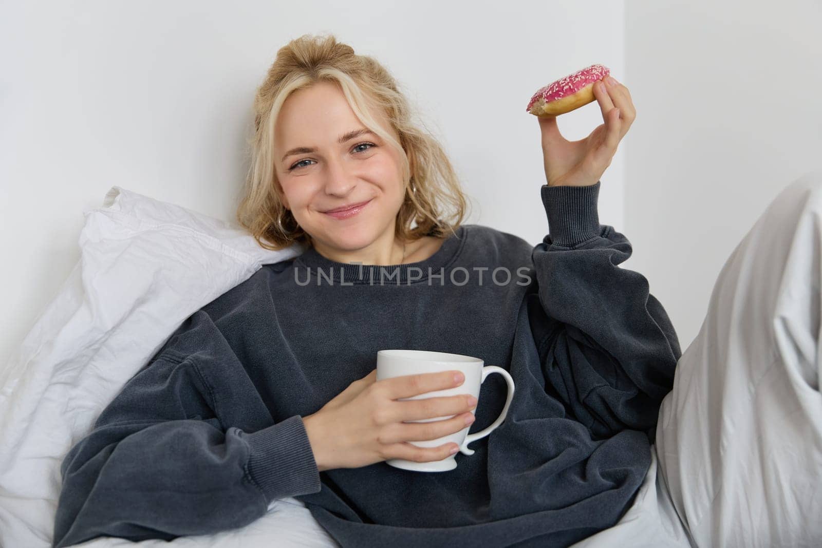 Lifestyle and people concept. Happy blond woman lying in bed with food, drinking tea and eating doughnut, resting at home, spending time alone, watching tv show.