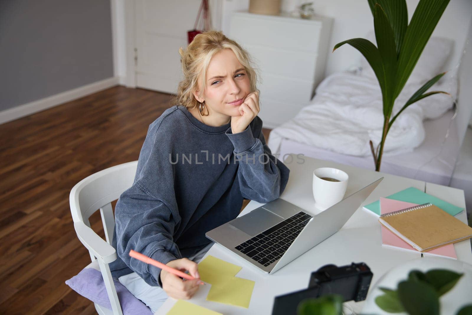 Portrait of young thoughtful girl, studying, making notes, writing down something in notebook, doing homework in her room, sitting in front of laptop, frowning while thinking.
