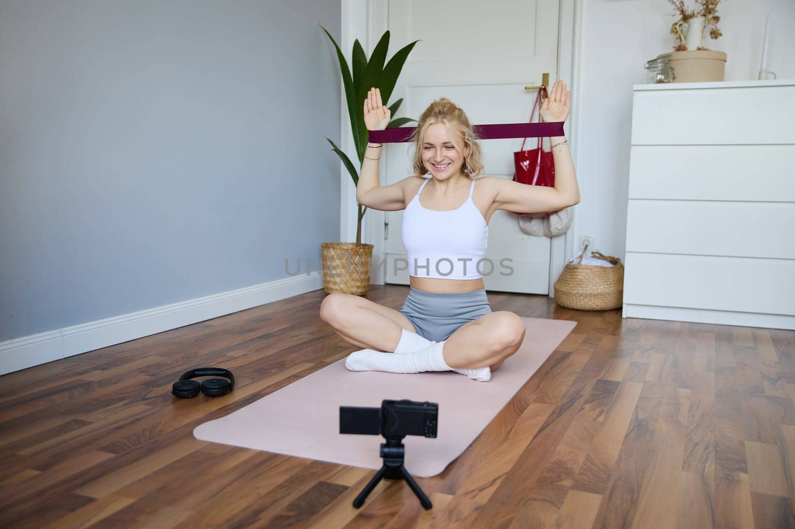 Portrait of young athletic woman recording home workout video, shooting content for sport fitness vlog, using resistance band and digital camera.