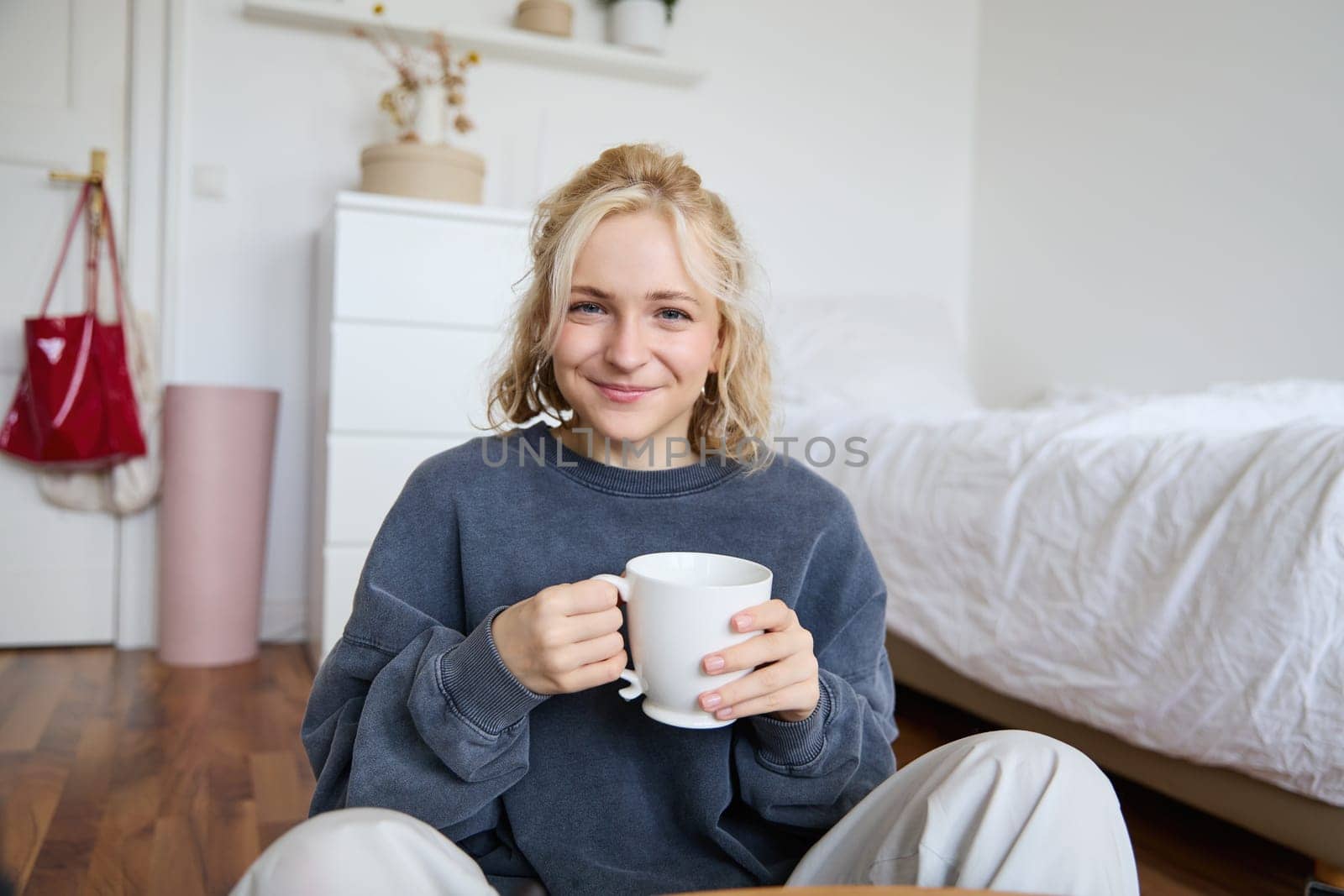 Portrait of young woman sitting on bedroom floor, drinking tea, holding white mug and smiling at camera.