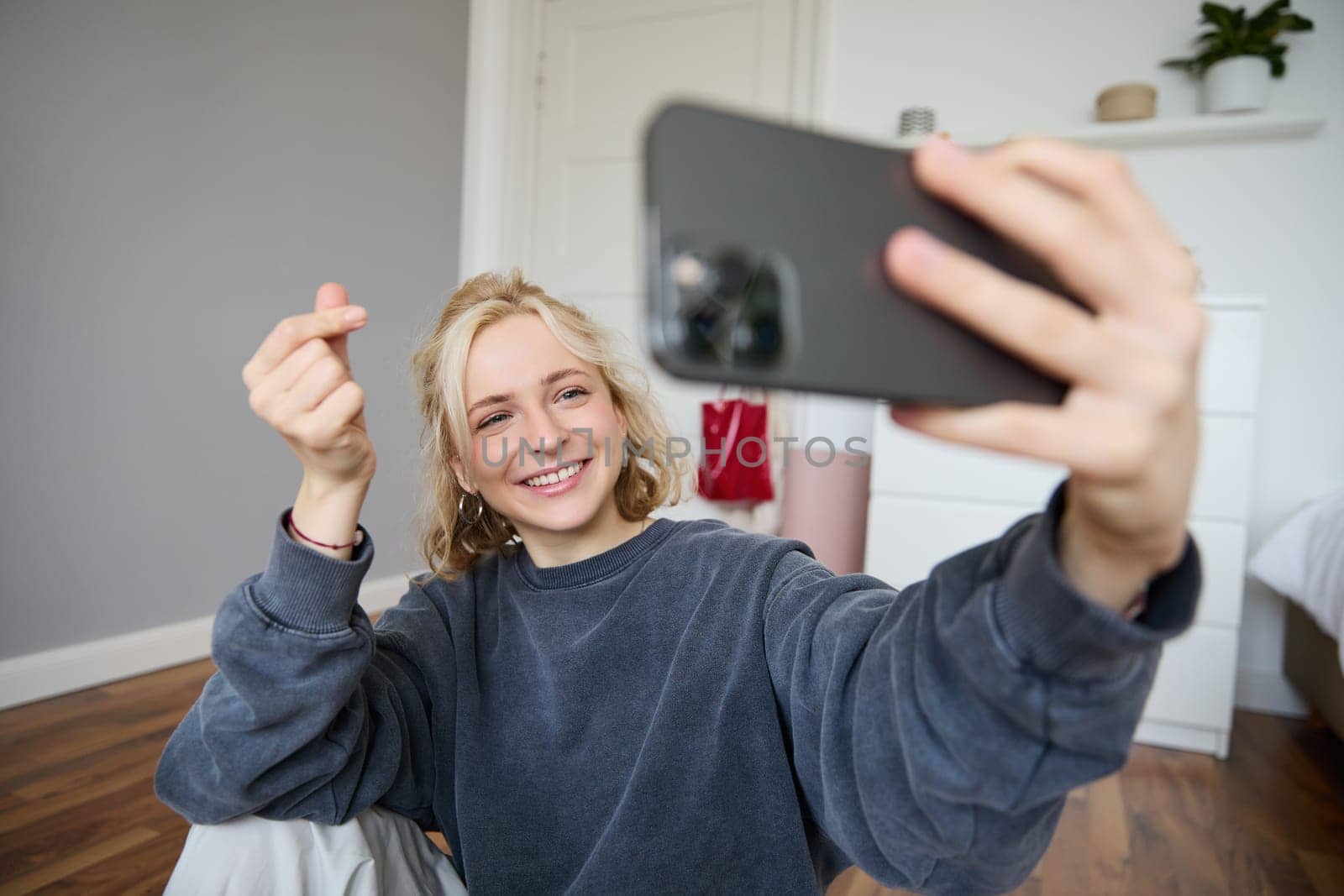 Image of young woman, vlogger taking selfie in her room, talking to her followers during online live stream, using smartphone app to chat with audience, smiling and looking happy.