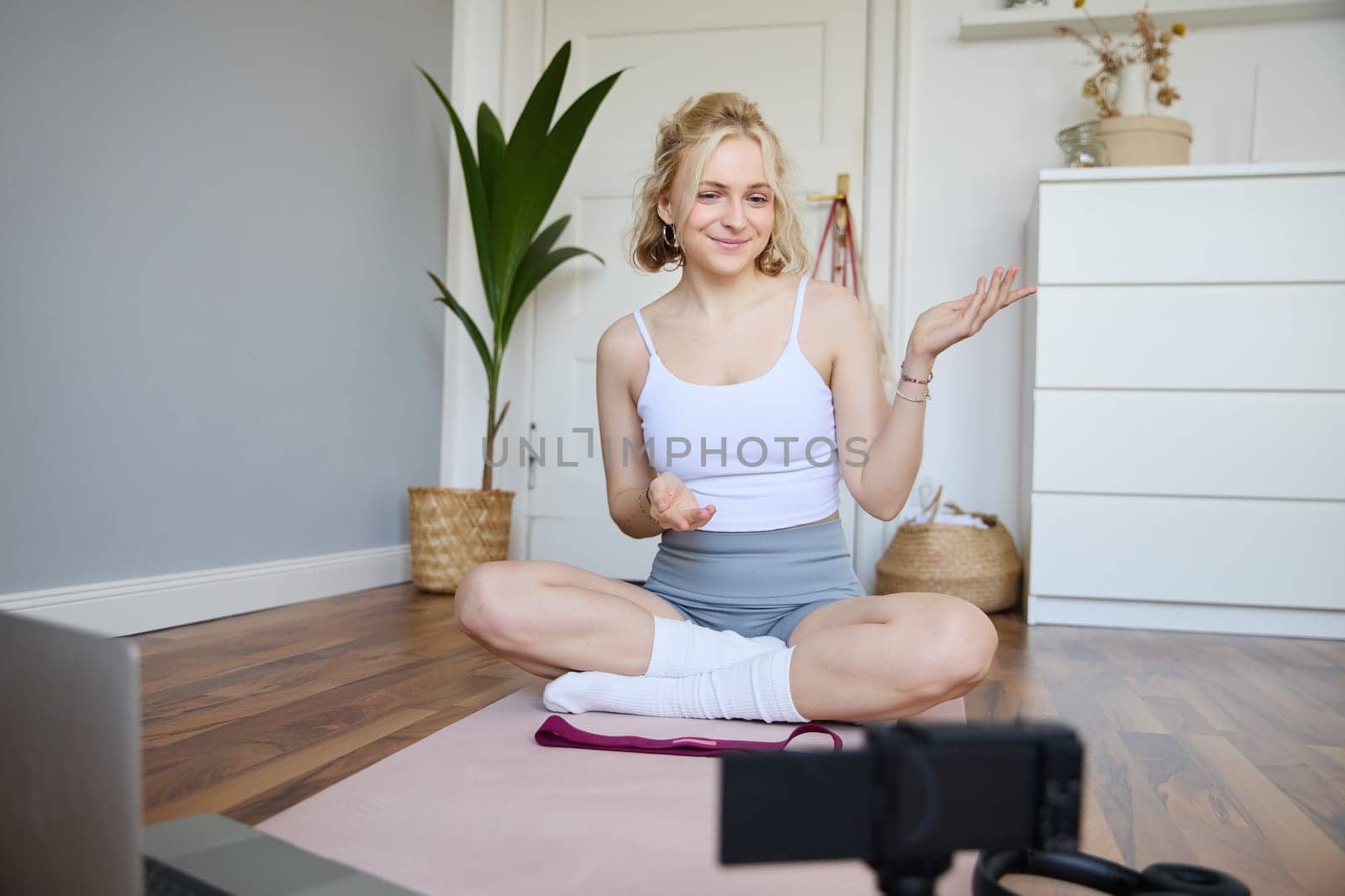 Portrait of beautiful fitness blogger, recording video on digital camera, showing workout exercises, explaining fitness movements or yoga training to followers, sits on rubber mat.