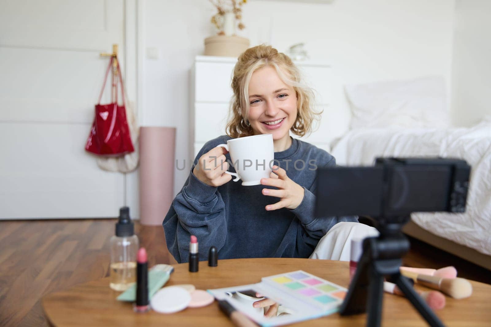 Cheerful woman, beauty blogger, records lifestyle vlog on digital camera, talks casually, tells a story for social media followers, holds cup, drinks tea and sits on floor in her room.