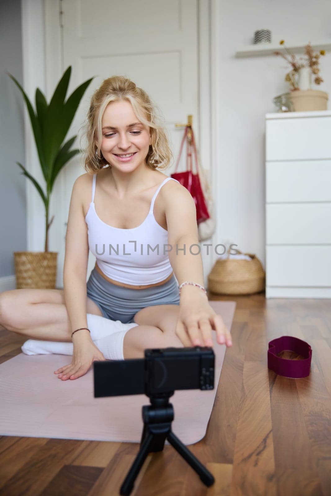 Vertical shot of woman recording her workout on digital camera, making a video about yoga and fitness at home, sitting on rubber mat in activewear.