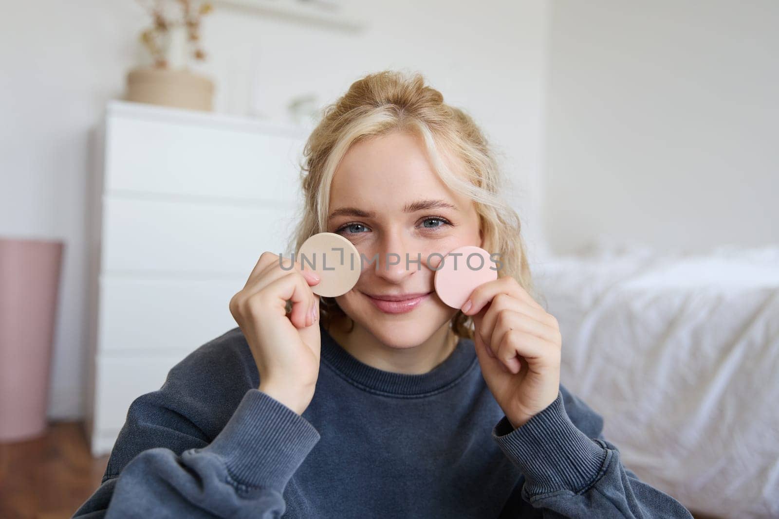 Image of young blond woman, girl records video about makeup, shows skin tone beauty products, sits in a room on floor.