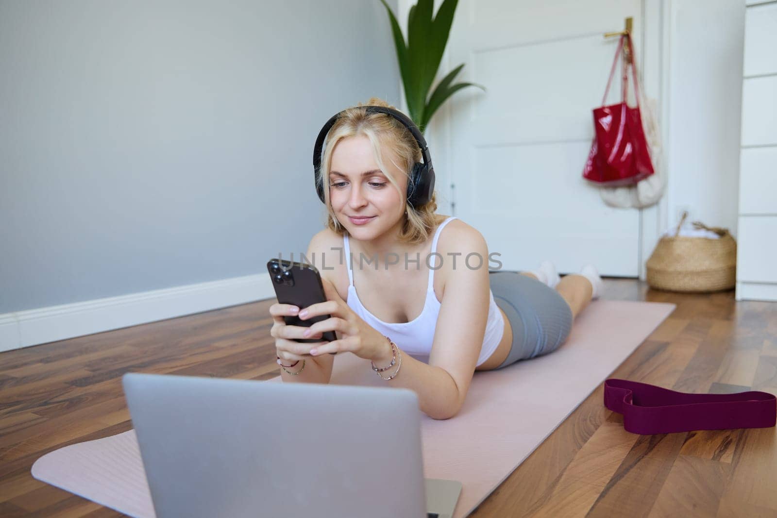 Portrait of young woman workout, watching exercise videos on laptop in headphones, lying on rubber mat with mobile phone and smiling.