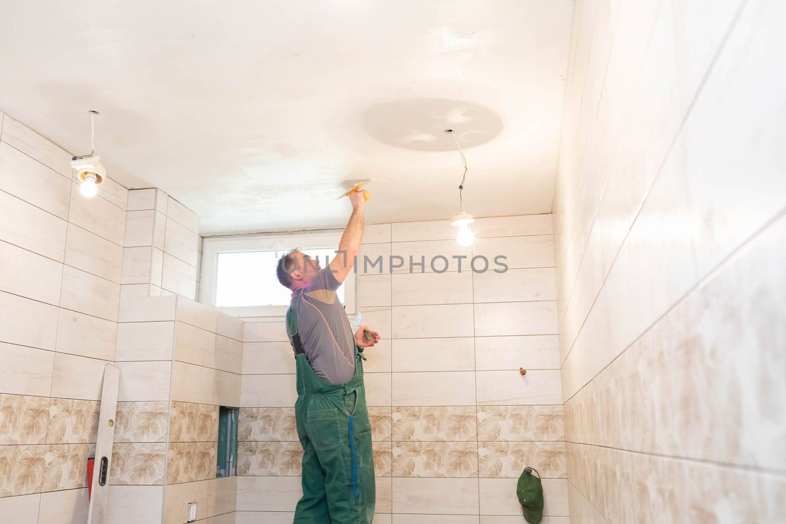 The worker applies plaster on the ceiling of the renovated bathroom. Construction worker in middle age. Green work clothes.