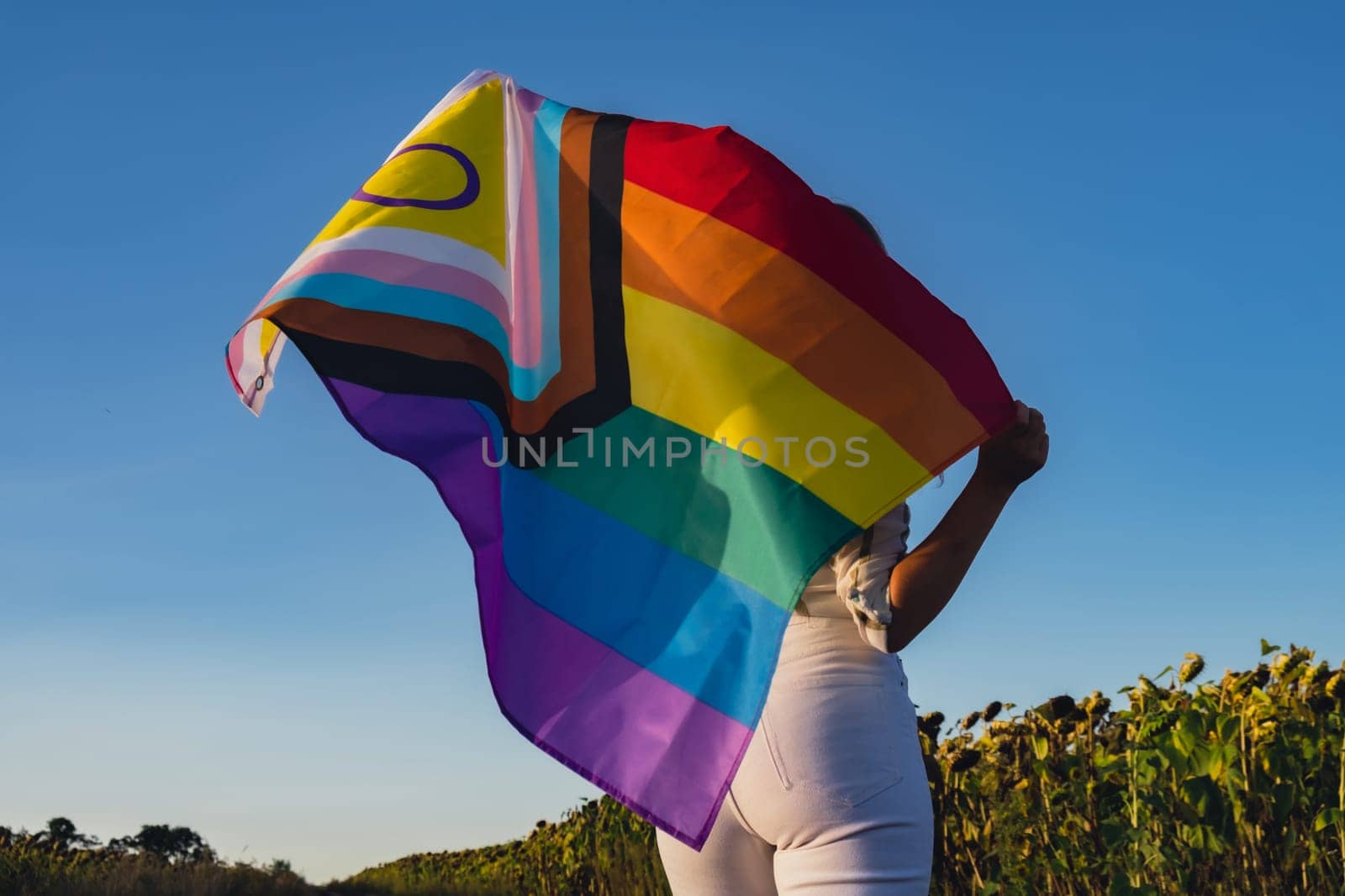 Girl showing her gender identity. Young woman stand with pride flag Rainbow LGBTQIA flag made from silk material on field background. Symbol of LGBTQ pride month. Equal rights. Peace and freedom concept