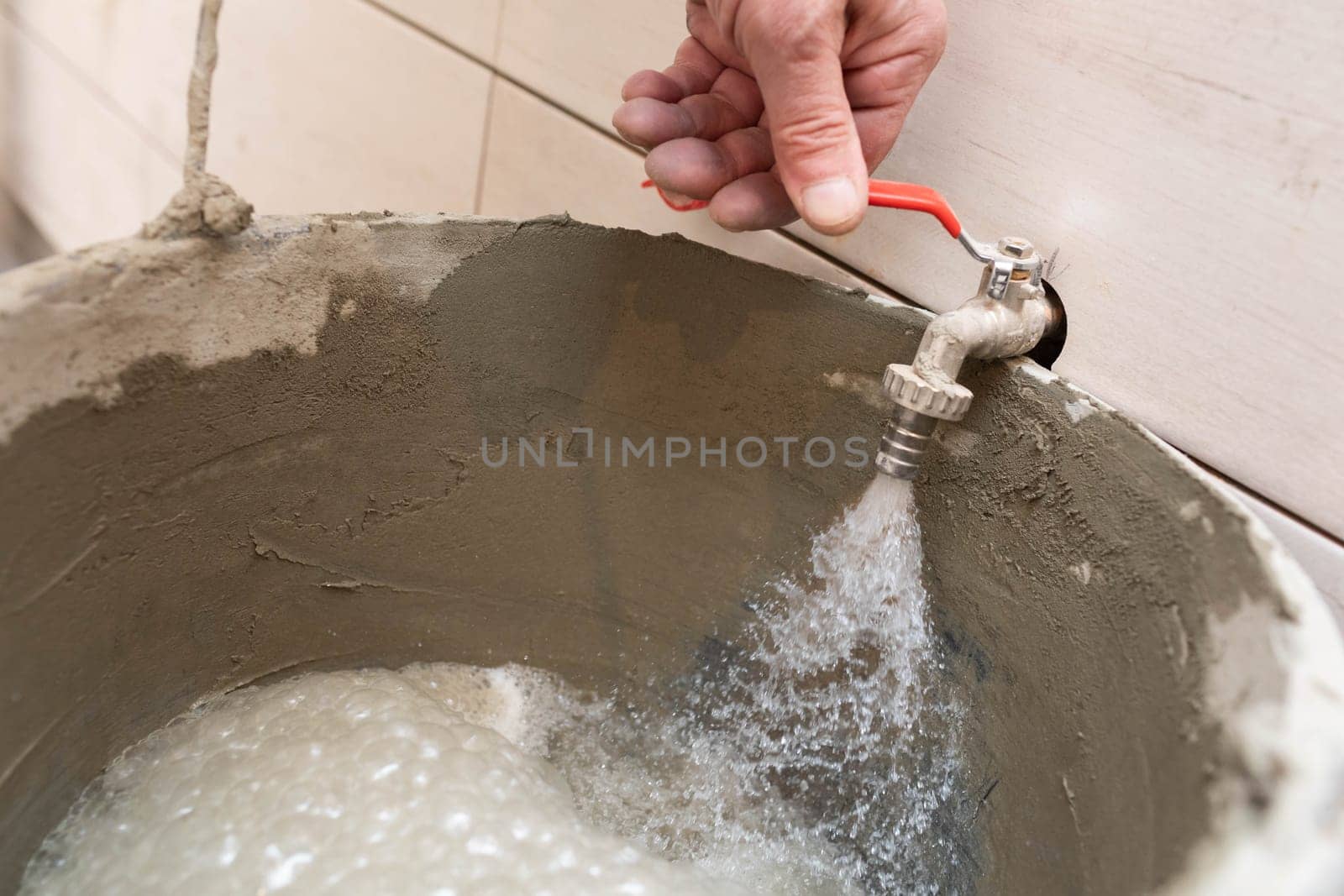 Construction worker. Unscrewed tap. Tap water flows into the bucket. During renovation. Tiles on the bathroom wall.