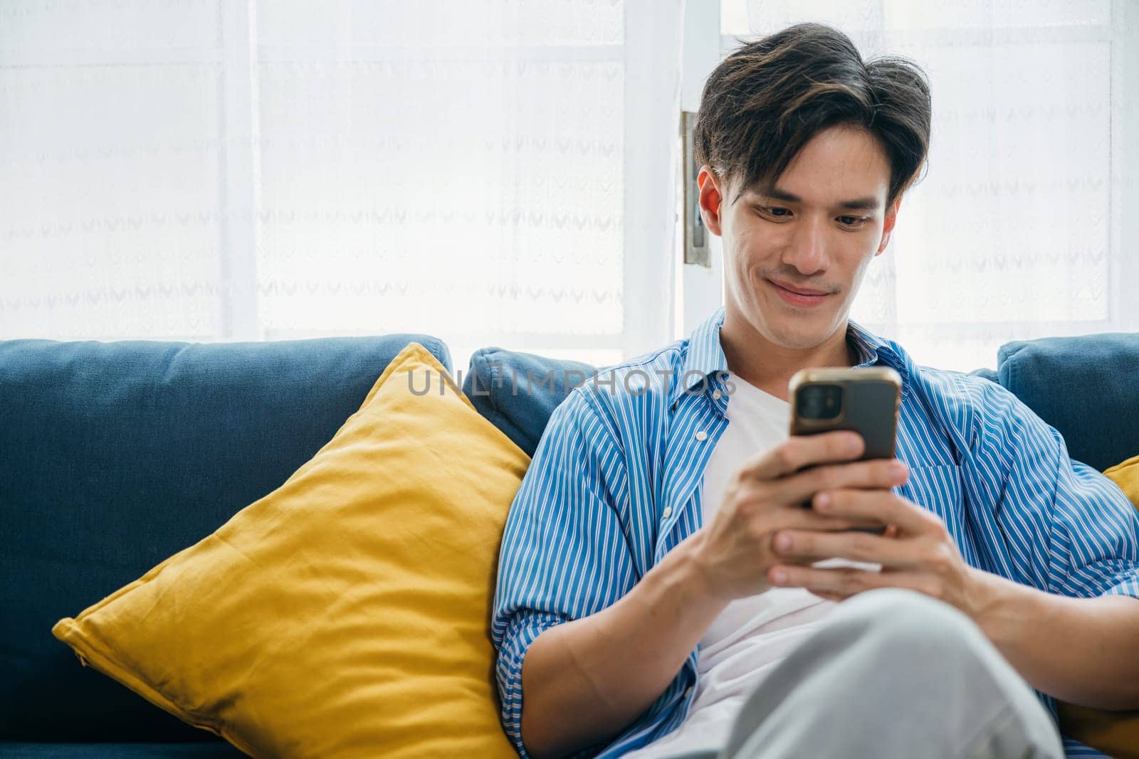 Engaged in a phone conversation a happy young man sits on a sofa enjoying relaxation. Embracing technology and communication he exudes happiness and success. scrolling on social media. by Sorapop