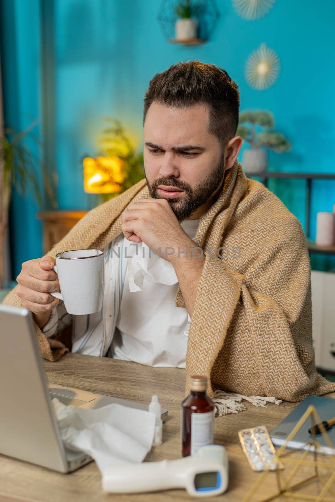 Sick ill young businessman suffering from cold flu or allergy working on laptop at home office. Manager freelancer man wrapped in blanket blows sneezes wipes snot into napkin. Coronavirus quarantine.