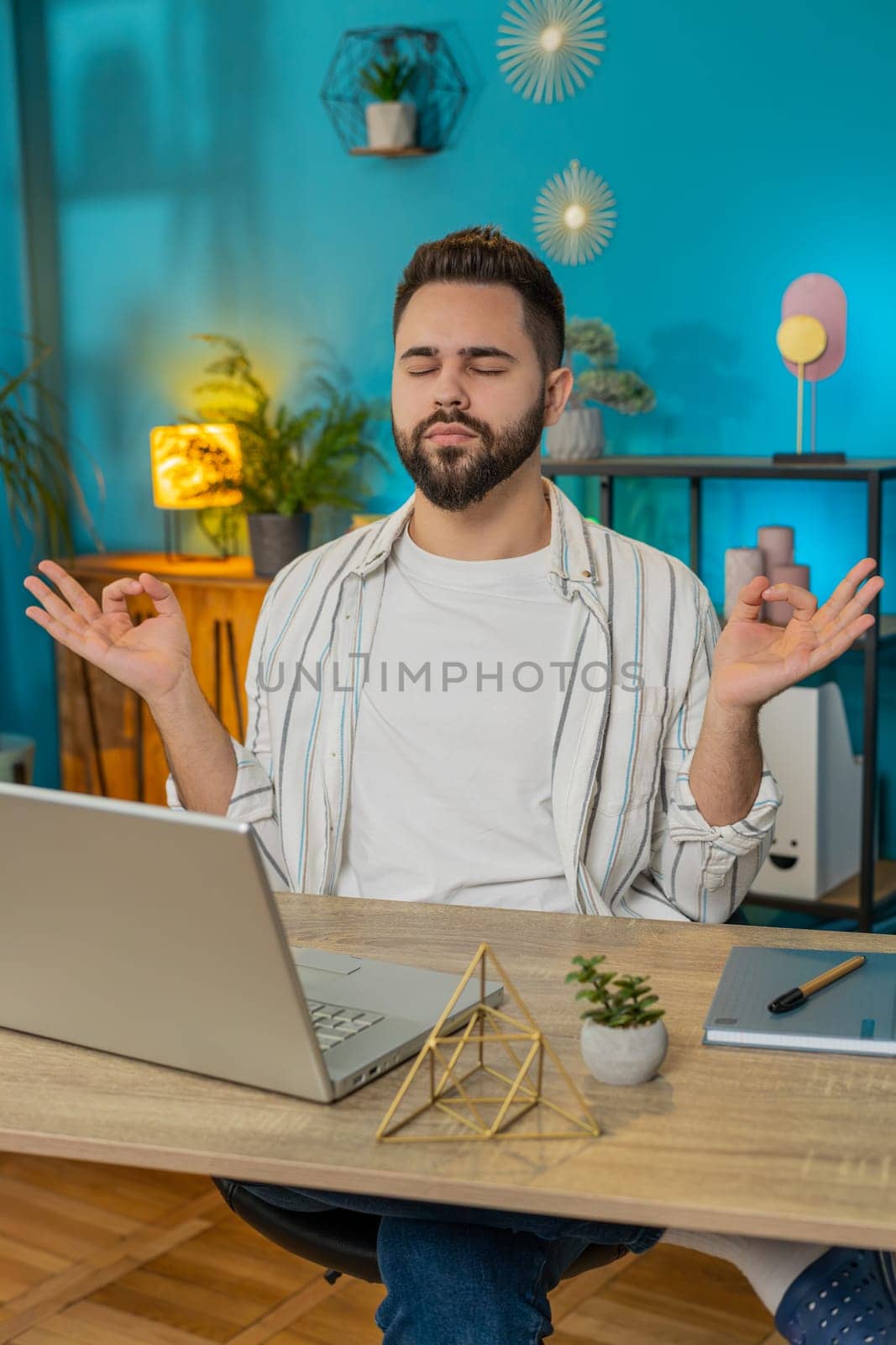 Peaceful relaxed male worker businessman sitting at workplace with folded in mudra gesture hands, reducing stress during workday, meditating or doing yoga breathing exercises in home office. Vertical