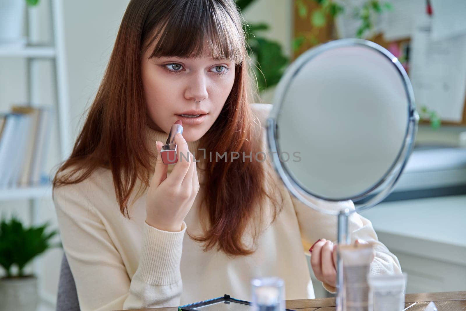 Young woman with cosmetic products on table, doing makeup looking in mirror at home, painting lips with lipstick. Cosmetics, fashion, youth, lifestyle, beauty concept