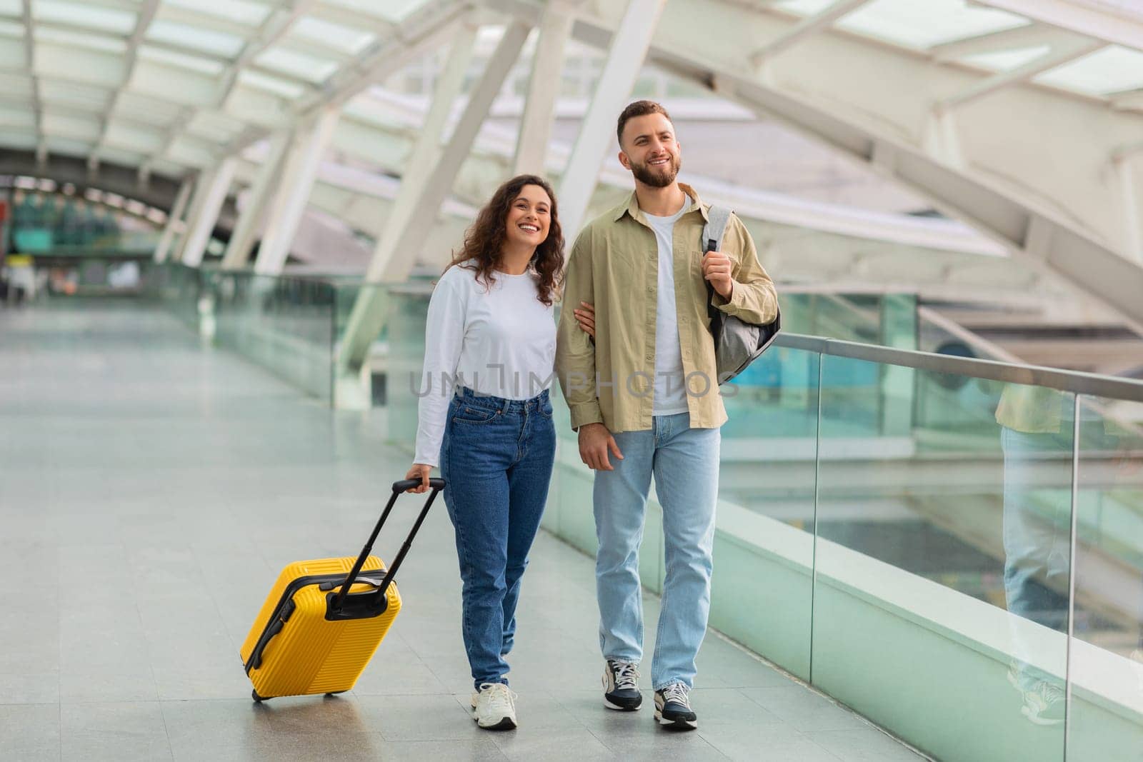Portrait Of Positive Young Couple Walking At Airport With Luggage, Happy Millennial Man And Woman Carrying Suitcase And Backpack Passing Hallway At Terminal, Enjoying Travelling Together