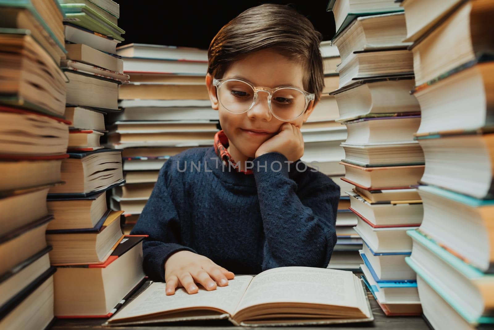 Cute little pupil boy in glasses reading book in library between stacks of books by kristina_kokhanova