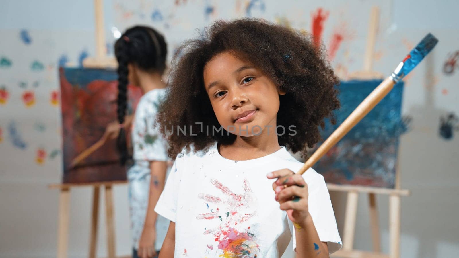 Cute girl holding painted brush while student drawing canvas behind. Erudition. by biancoblue