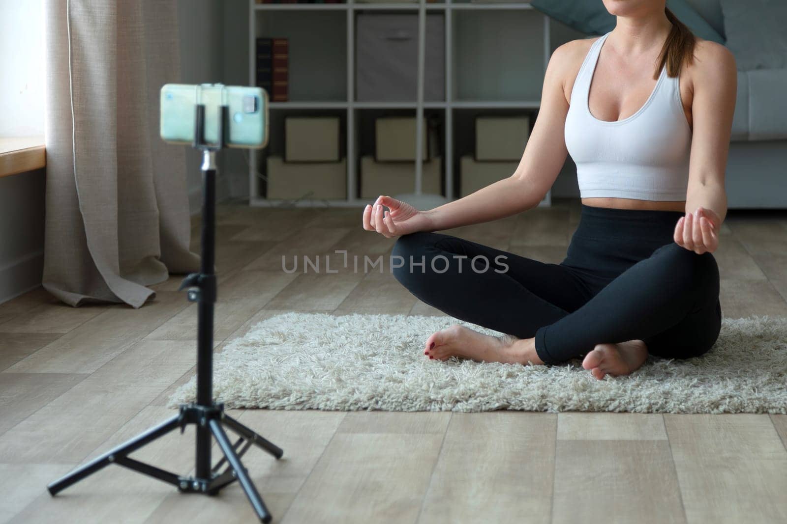 woman doing yoga at home on the carpet, sitting in lotus position with her phone and tripod for recording videos or live streaming for an online fitness center class through a social media platform. She is sitting in the style of lotus position