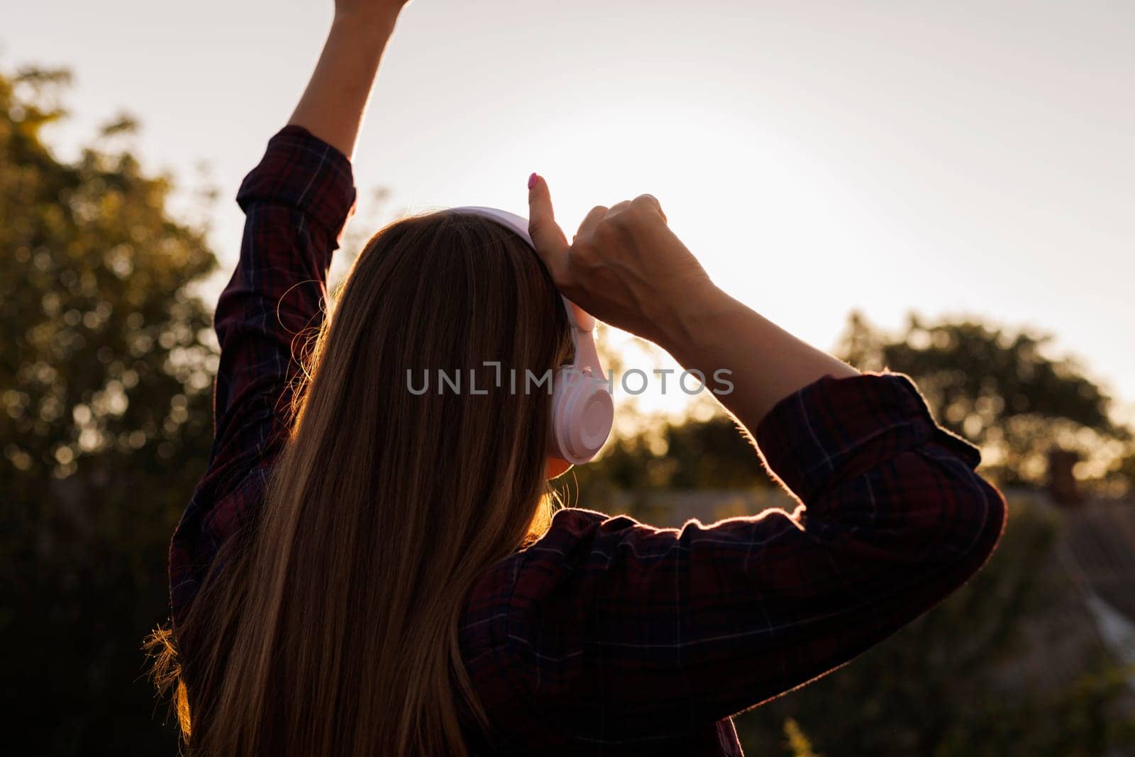Back view of woman with headphones raising arms against sunset. Outdoor music enjoyment concept with copy space for design and print