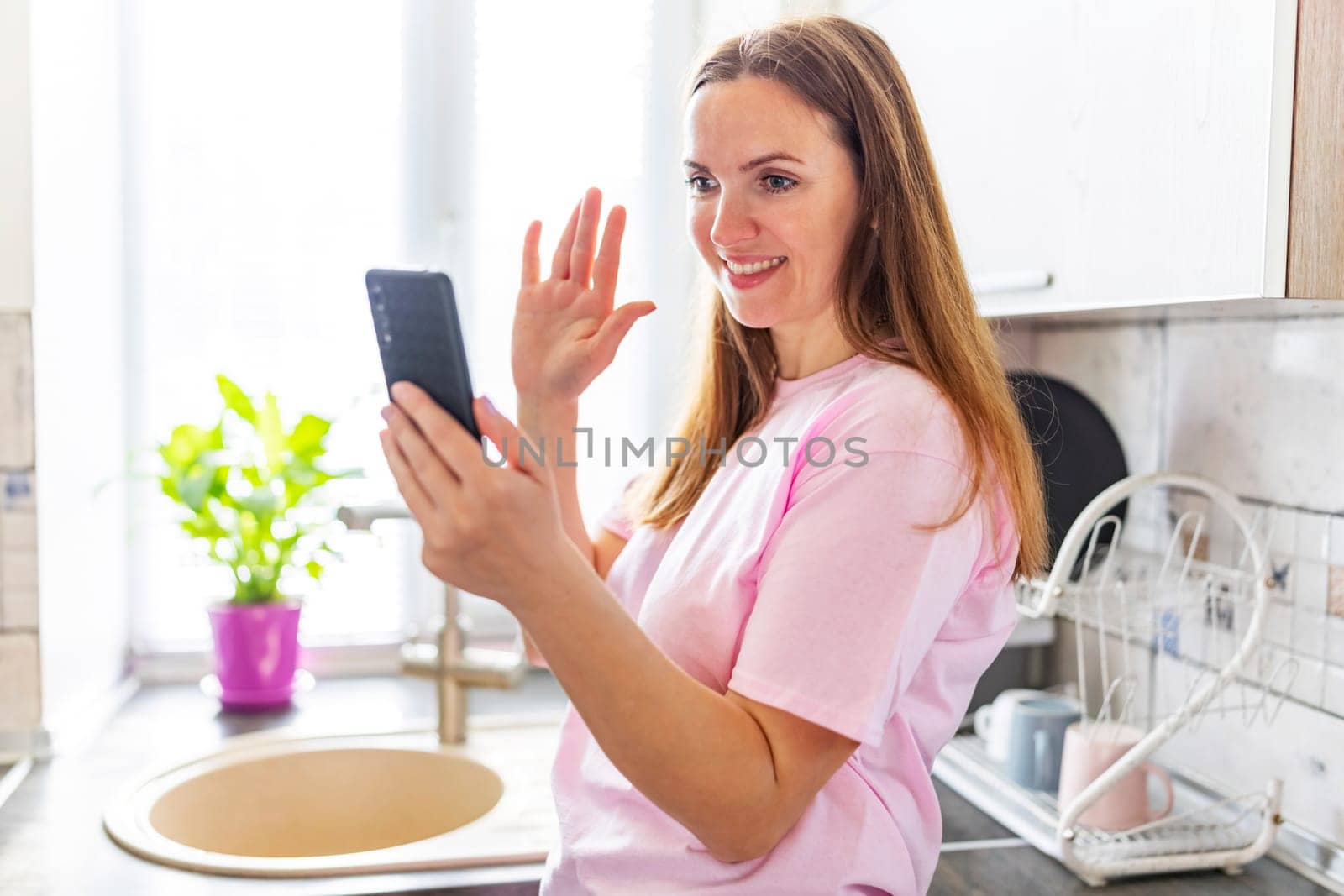 Woman waving at phone during a video call in a bright kitchen. Casual home lifestyle concept