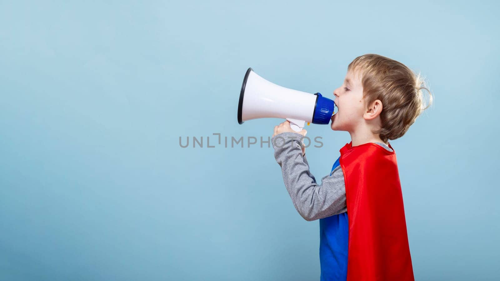 Child in superhero costume using megaphone. Blue background with copy space. Childhood imagination and play concept. Design for announcements, children's events