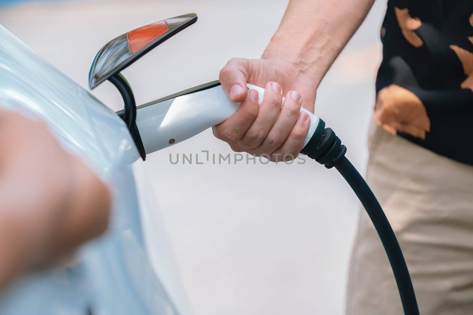 Hand insert EV charger plug into electric vehicle to recharge EV car battery from outdoor charging station. Fresh daylight environment with alternative clean and sustainable energy concept. Perpetual