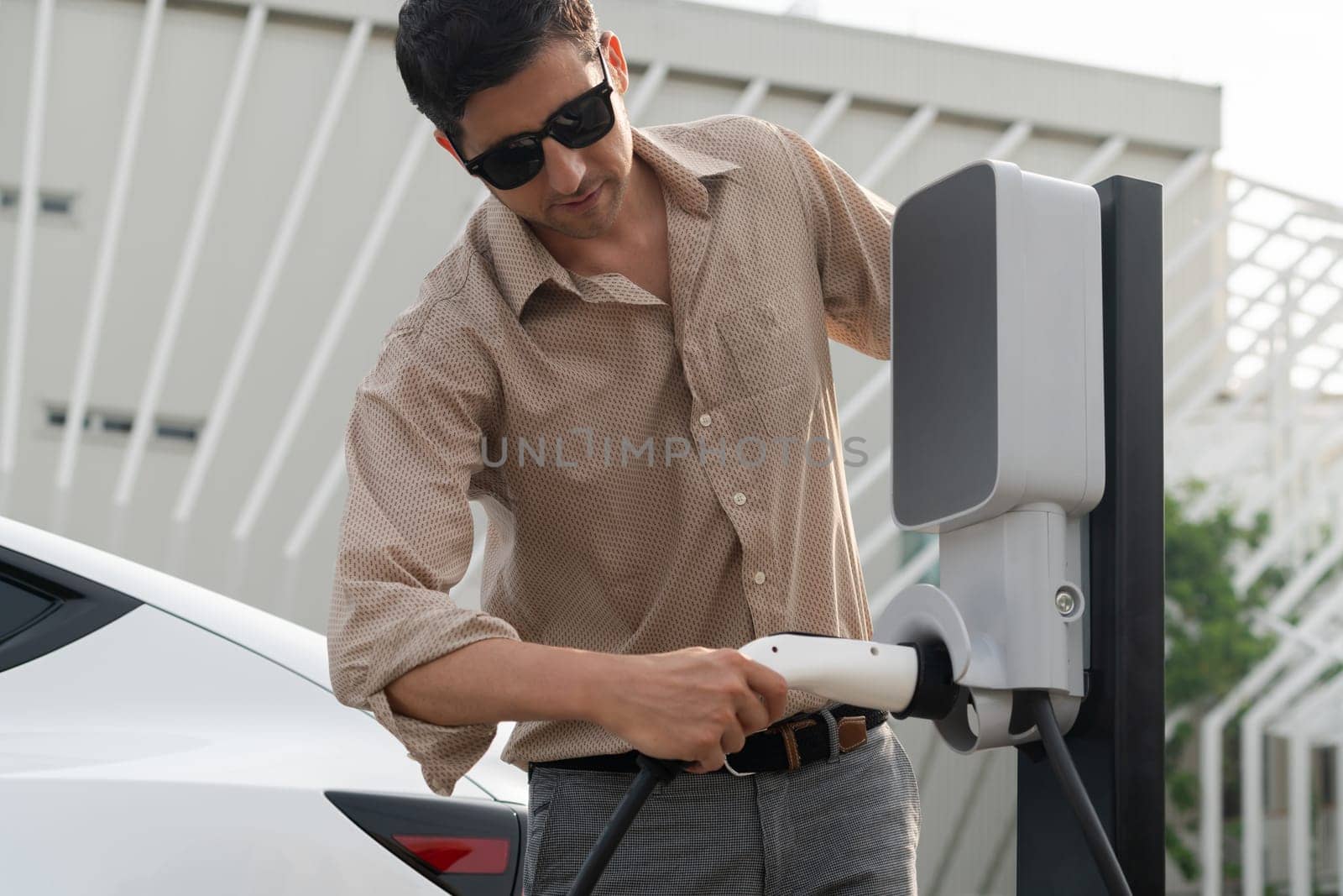 Young man put EV charger to recharge electric car's battery from charging station in city commercial parking lot. Rechargeable EV car for sustainable environmental friendly urban travel. Expedient