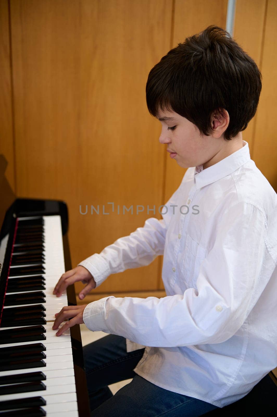 Confident portrait of teen boy in white shirt, musician pianist putting fingers on piano keys while playing piano, enjoying the performance of classical music indoor. Kids education and entertainment.