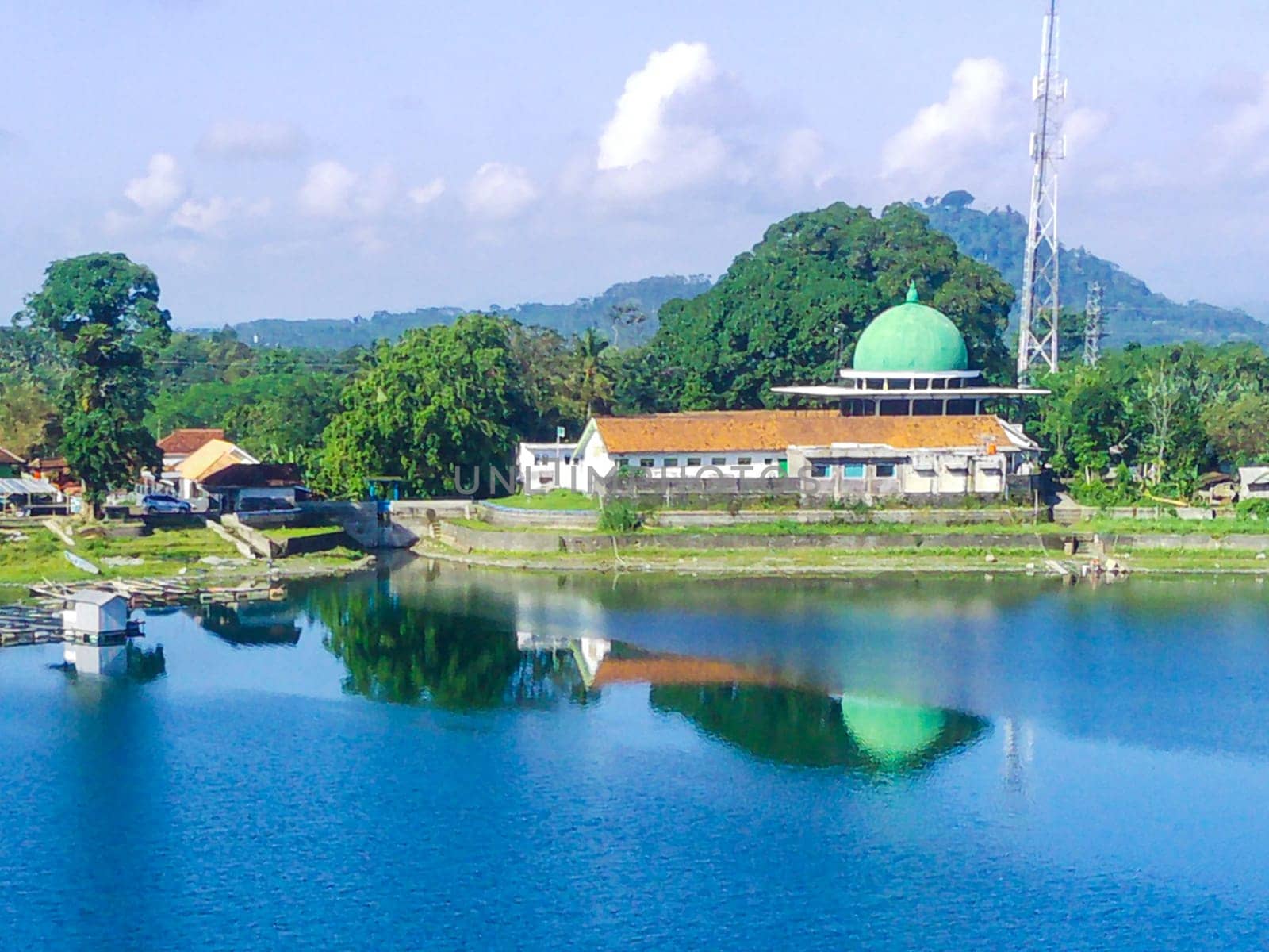 serene lake surrounded by lush green trees, with a white building with a mosque in the background. Around Ranu or Lake Klakah, Lumajang, East Java, Indonesia.