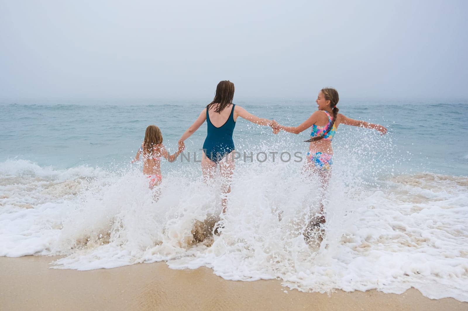 Mother and children playing on the ocean beach. Family enjoying the ocean. Mother holds girls's hands and they all look at the ocean together by esvetleishaya