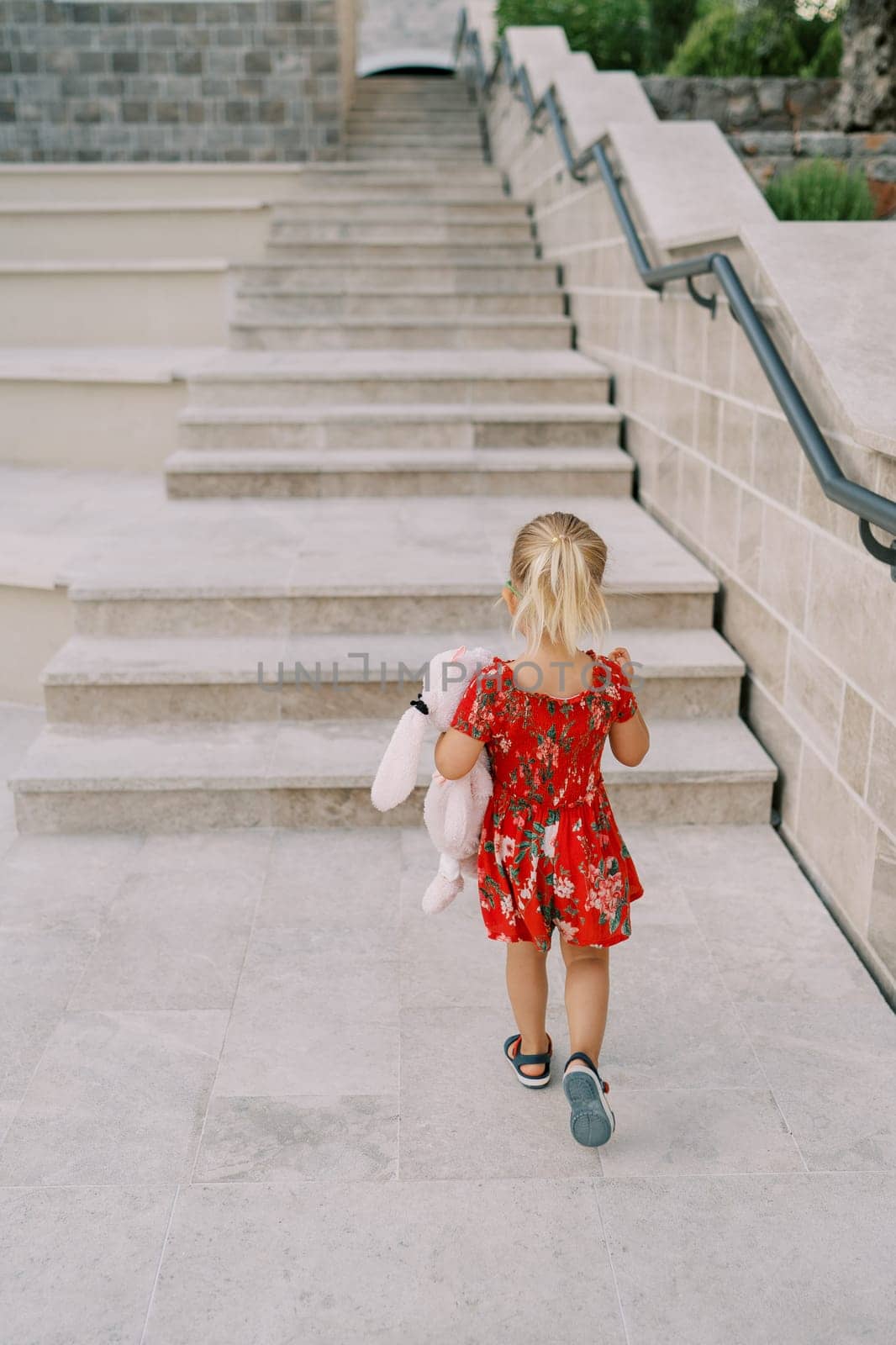 Little girl with a pink toy hare under her arm walks up the high steps of a building. Back view. High quality photo