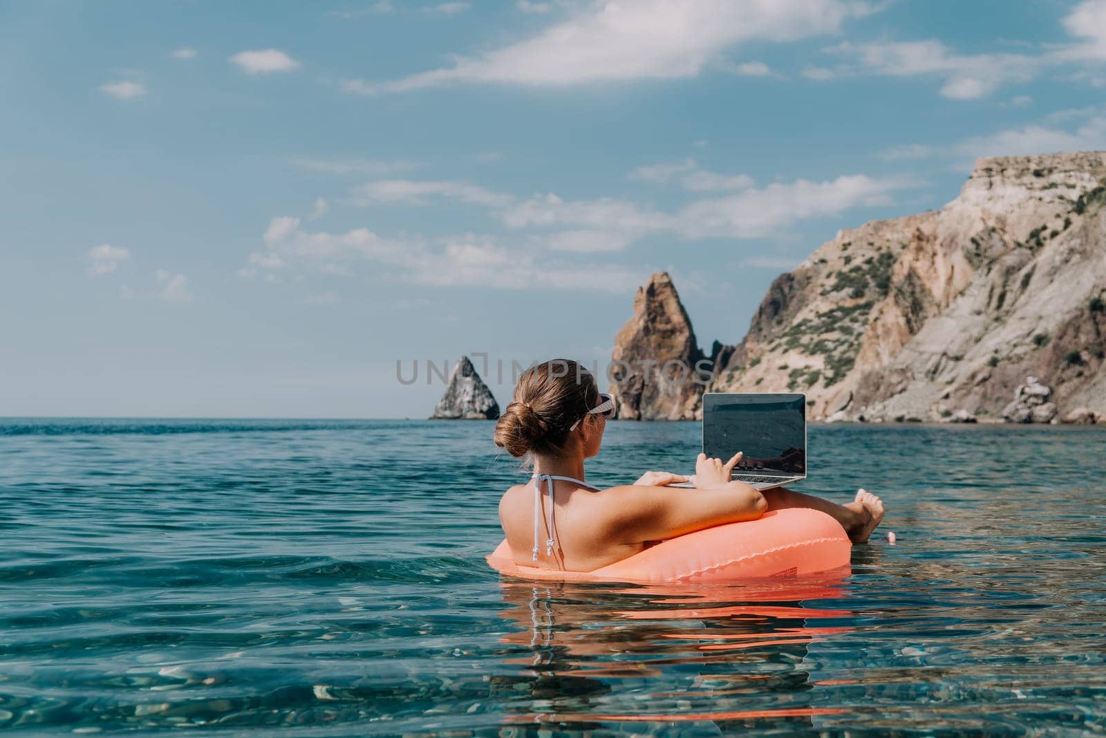 Woman freelancer works on laptop swimming in sea on pink inflatable ring. Pretty lady typing on computer while floating in the sea on inflatable donut at sunset. Freelance, remote work on vacation