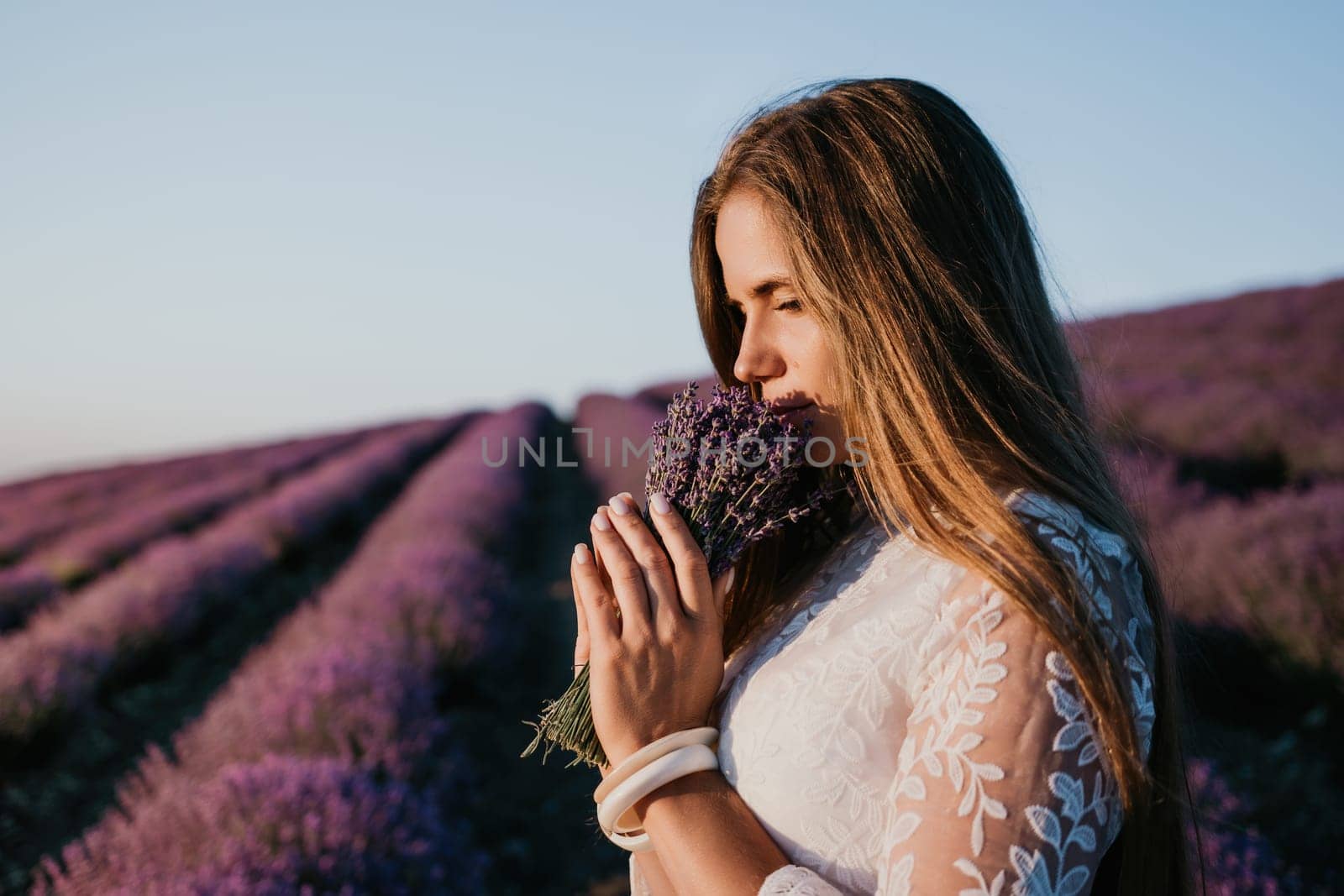 Close up portrait of young beautiful woman in a white dress and a hat is walking in the lavender field and smelling lavender bouquet.